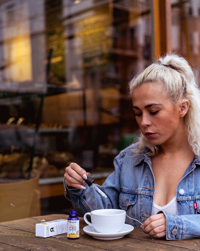 Blonde woman in denim jacket mixing Binoid CBD oil into a coffee cup at an outdoor café.